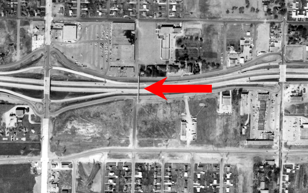 A top-down black and white image taken from a plane that shows the area around the pedestrian bridge, which is marked with a large red arrow.  Crossing the interestate on either side of the pedestrian bridge are two other bridges for cars, on the north we can see some buildings and parking lots, and on the south there is an empty lot.  Far north and far south we can see houses.