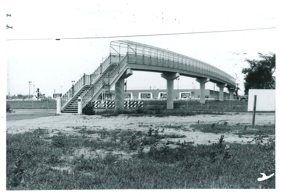 Black-and-white photo of the bridge taken from the ground at an angle.  In the foreground is some grass and sand; the photographer is standing on an empty lot.