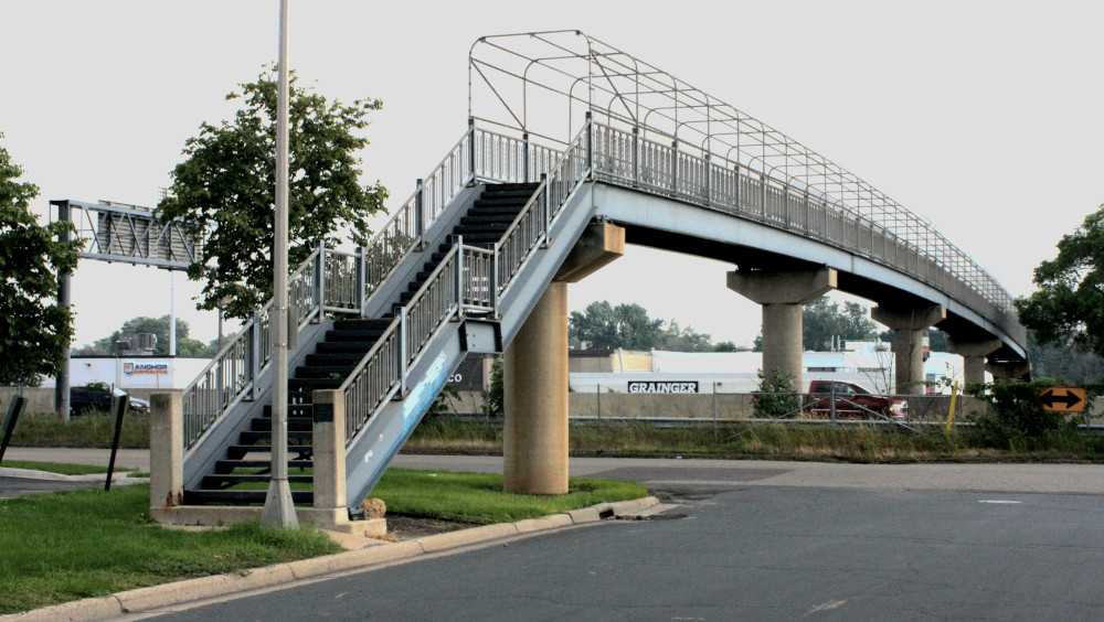 Photo of the base of the pedestrian bridge showing stairs that lead from a grassy median up to the bridge over a road and then over the interstate.